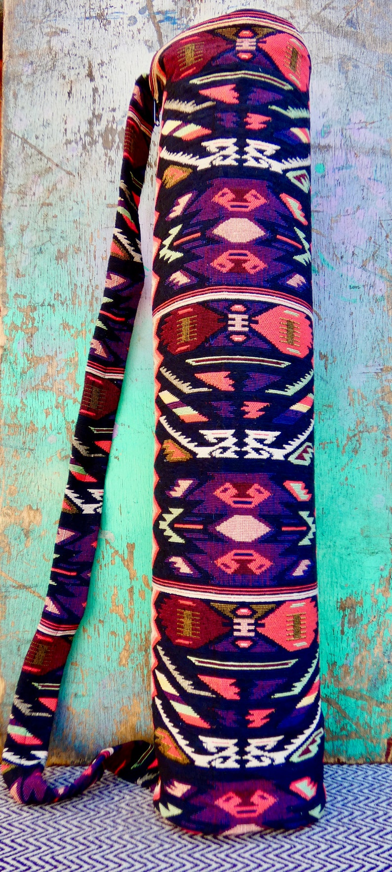 May include: A black yoga mat bag with a colorful geometric pattern in shades of pink, red, purple, white, and gold. The bag has a long strap for carrying over the shoulder.