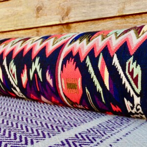 May include: A long, cylindrical pillow with a colorful, geometric pattern in shades of pink, blue, green, and gold. The pillow is made of a woven fabric and has a fringed edge.