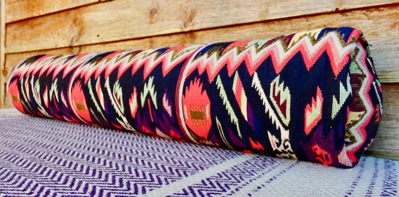 May include: A long, cylindrical pillow with a colorful, geometric pattern in shades of pink, blue, green, and gold. The pillow is made of a woven fabric and has a fringed edge.