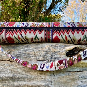 Pu&ograve; includere: Una borsa beige per tappetino da yoga con un motivo geometrico colorato in rosso, verde e viola. La borsa ha una cerniera nera e una tracolla fantasia abbinata. La borsa &egrave; appoggiata su una superficie di legno stagionato.