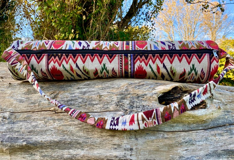 Pu&ograve; includere: Una borsa beige per tappetino da yoga con un motivo geometrico colorato in rosso, verde e viola. La borsa ha una cerniera nera e una tracolla fantasia abbinata. La borsa &egrave; appoggiata su una superficie di legno stagionato.