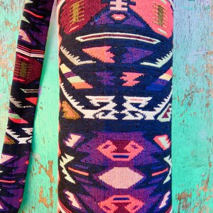 May include: A yoga mat bag with a colorful geometric pattern in shades of pink, purple, black, white, and gold. The bag is made of a woven fabric and has a strap.