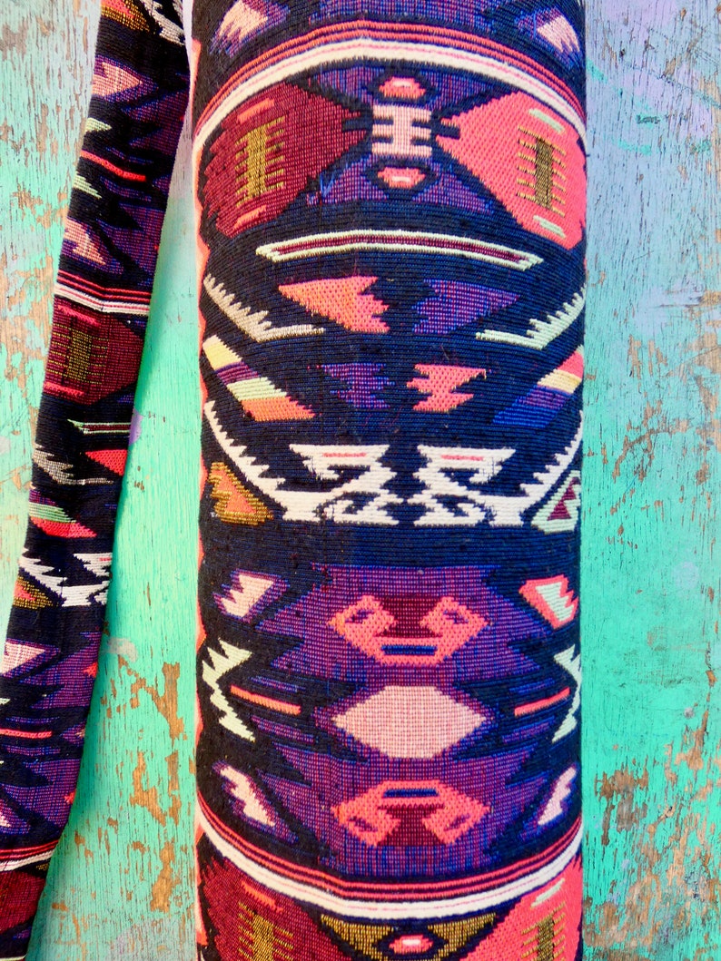 May include: A yoga mat bag with a colorful geometric pattern in shades of pink, purple, black, white, and gold. The bag is made of a woven fabric and has a strap.