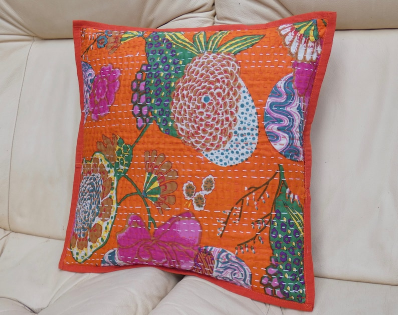 Peut inclure: Coussin carr&eacute; orange avec un motif floral vibrant en rose, vert et bleu. Le coussin a une bordure rouge et des coutures blanches. Il est pos&eacute; sur une surface blanche.