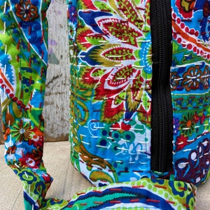 May include: A patterned bag with a black zip and a matching strap. The bag features a vibrant mix of blue, green, red, and white floral and paisley designs. The fabric appears to be quilted with white stitching.