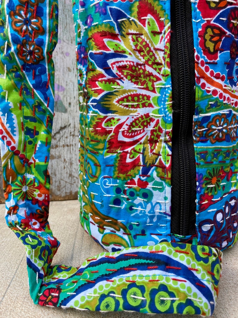 May include: A patterned bag with a black zip and a matching strap. The bag features a vibrant mix of blue, green, red, and white floral and paisley designs. The fabric appears to be quilted with white stitching.