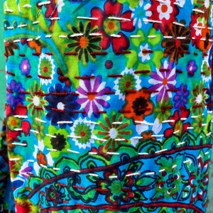May include: A close-up shot of a colourful textile with a floral design. The fabric is predominantly blue, with flowers in red, orange, purple, and green. White and brown stitching adds texture. The pattern includes both large and small floral elements.