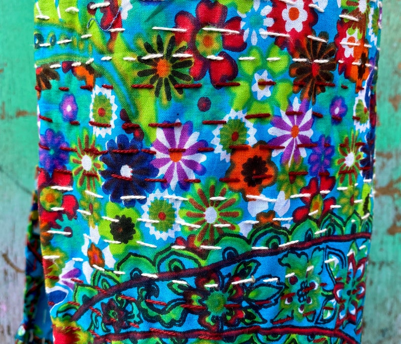 May include: A close-up shot of a colourful textile with a floral design. The fabric is predominantly blue, with flowers in red, orange, purple, and green. White and brown stitching adds texture. The pattern includes both large and small floral elements.