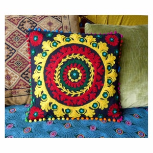 Puede incluir: Un coj&iacute;n decorativo cuadrado azul marino con un vibrante dise&ntilde;o floral bordado en rojo, amarillo y verde. El coj&iacute;n est&aacute; adornado con pompones de colores y forma parte de una colecci&oacute;n de decoraci&oacute;n del hogar.