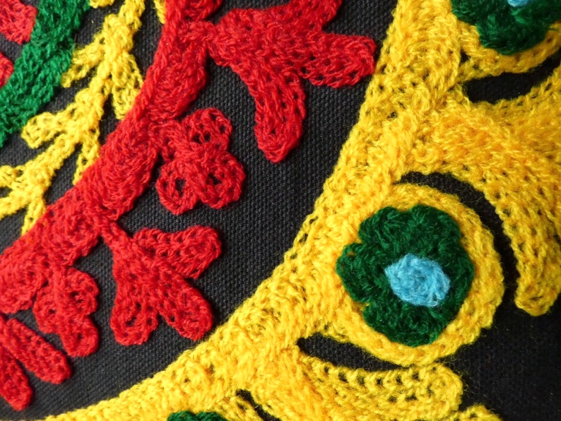 Puede incluir: Primer plano de una tela negra con vibrantes patrones florales bordados. El bordado presenta hilos rojos, amarillos y verdes, creando un dise&ntilde;o decorativo texturizado. El dise&ntilde;o incluye hojas estilizadas y formas de flores.