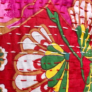 K&ouml;nnte beinhalten: Nahaufnahme eines roten Stoffes mit wei&szlig;en, gr&uuml;nen und gelben Blumenstickereien. Der Stoff hat eine gesteppte Textur mit wei&szlig;en N&auml;hten.