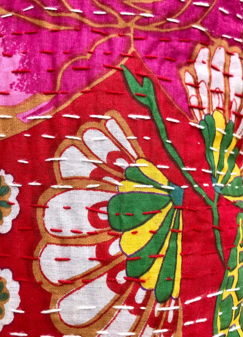 K&ouml;nnte beinhalten: Nahaufnahme eines roten Stoffes mit wei&szlig;en, gr&uuml;nen und gelben Blumenstickereien. Der Stoff hat eine gesteppte Textur mit wei&szlig;en N&auml;hten.