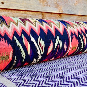 May include: A long, cylindrical pillow with a colorful geometric pattern in shades of pink, blue, green, and gold. The pillow is made of a woven fabric and has a fringed edge.