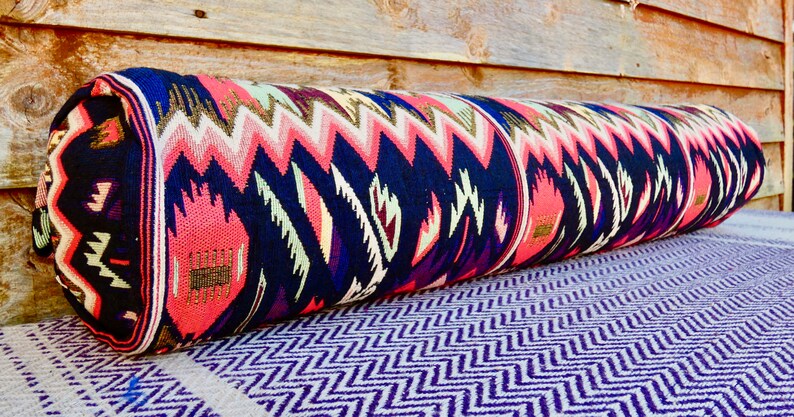 May include: A long, cylindrical pillow with a colorful geometric pattern in shades of pink, blue, green, and gold. The pillow is made of a woven fabric and has a fringed edge.