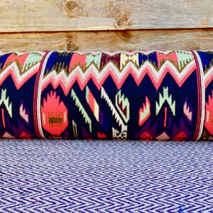 May include: A long, cylindrical pillow with a colorful geometric pattern in shades of pink, blue, and white. The pillow is made of fabric with a woven texture.