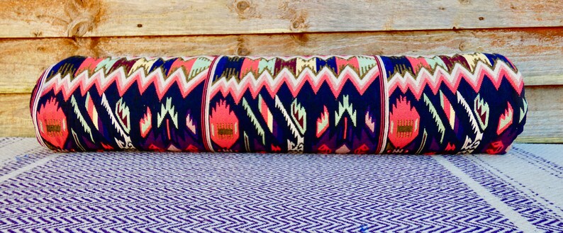 May include: A long, cylindrical pillow with a colorful geometric pattern in shades of pink, blue, and white. The pillow is made of fabric with a woven texture.