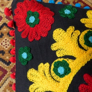 Puede incluir: Un coj&iacute;n negro decorativo con vibrantes dise&ntilde;os florales bordados en rojo, amarillo y verde. El coj&iacute;n presenta un borde de pompones rojos y azules, a&ntilde;adiendo un toque l&uacute;dico al textil artesanal.