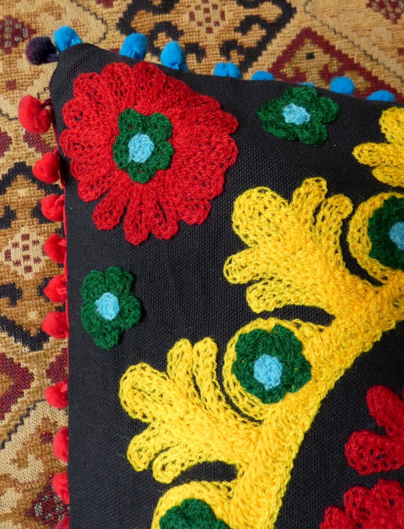 Puede incluir: Un coj&iacute;n negro decorativo con vibrantes dise&ntilde;os florales bordados en rojo, amarillo y verde. El coj&iacute;n presenta un borde de pompones rojos y azules, a&ntilde;adiendo un toque l&uacute;dico al textil artesanal.