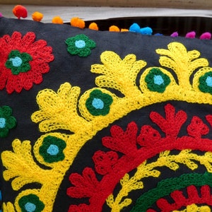 Puede incluir: Un coj&iacute;n negro decorativo con vibrantes bordados florales en rojo, amarillo, verde y azul. El coj&iacute;n presenta un ribete de pomp&oacute;n en varios colores. El dise&ntilde;o es intrincado y detallado, adecuado para la decoraci&oacute;n del hogar.