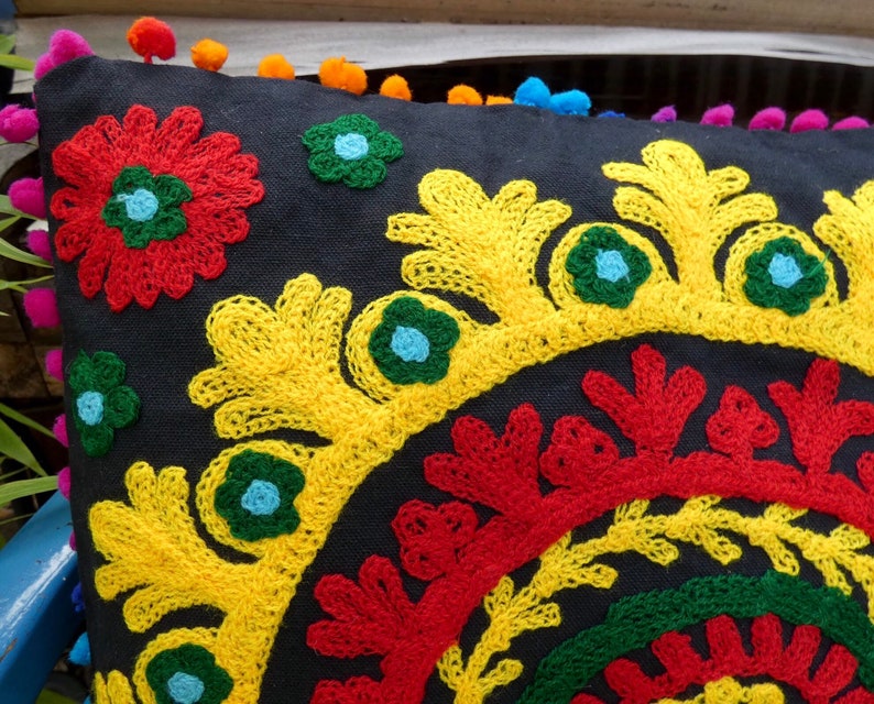 Puede incluir: Un coj&iacute;n negro decorativo con vibrantes bordados florales en rojo, amarillo, verde y azul. El coj&iacute;n presenta un ribete de pomp&oacute;n en varios colores. El dise&ntilde;o es intrincado y detallado, adecuado para la decoraci&oacute;n del hogar.
