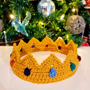 May include: A handcrafted, golden-yellow crochet crown with a zig-zag edge. The crown features colourful, jewel-like accents in blue, purple, red, and green. The crown is likely for a child or pet, set against a blurred background.