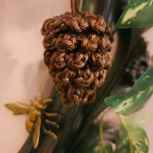 May include: A brown crocheted pine cone ornament hanging from a branch with green leaves.
