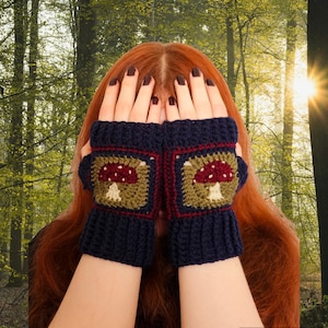 May include: Dark blue crochet fingerless gloves with a square mushroom design in the center. The mushroom has a red cap and a cream-colored stem, set against a green and brown background. The gloves are displayed against a backdrop of a forest.