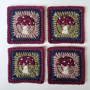 May include: Four square crochet coasters with a mushroom design. Each coaster features a burgundy border, with concentric rings of blue, olive green, and tan yarn. The mushroom is burgundy with white spots, and a white stem.