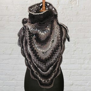 May include: A triangular crochet shawl featuring a complex, layered pattern in shades of black, charcoal, and light gray. The design incorporates intricate stitch work with scalloped edges and a textured, openwork construction throughout the piece.