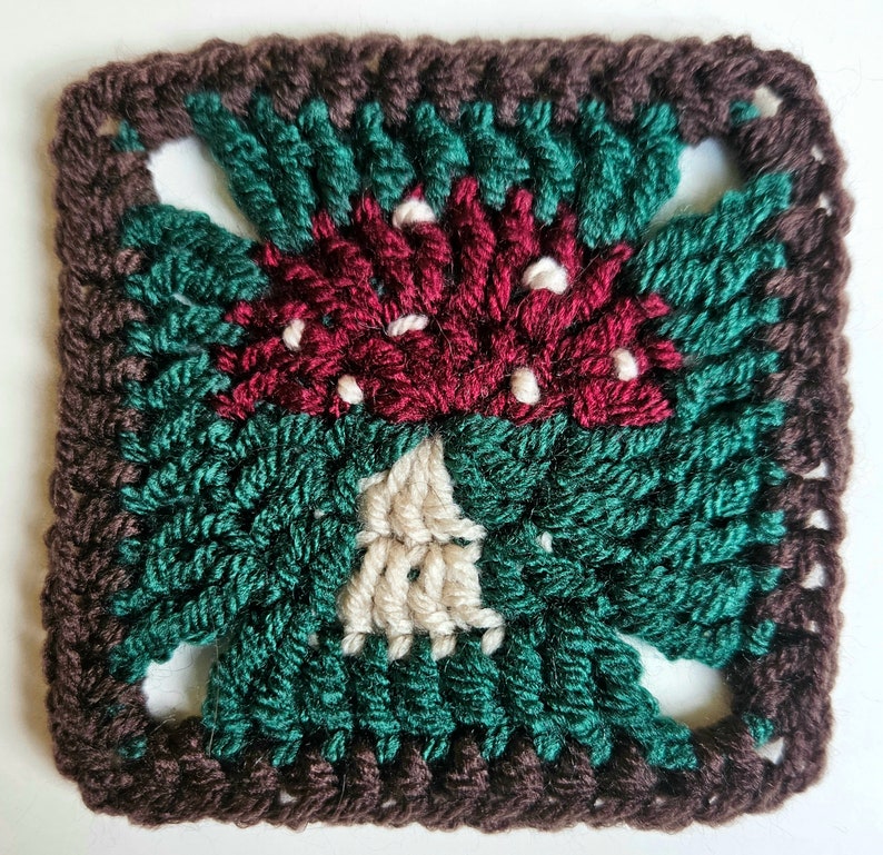 This image shows a singular crocheted square with a toadstool motif, shown on a plain white background. The toadstool is red and white, with white dots. It has a deep green surround and a brown border.