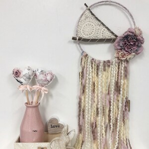 Può includere: Un acchiappasogni floreale rosa e bianco con un cerchio di legno e un triangolo all'uncinetto. L'acchiappasogni è appeso a un muro bianco e ha fili di lana rosa e bianco. Ci sono due cuori floreali rosa in un vaso rosa e un coniglio di legno su una superficie bianca. Il coniglio di legno ha la parola "Love" su di esso.