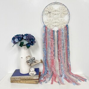 Può includere: Un acchiappasogni bianco all'uncinetto con nappe di tessuto blu e rosa che pendono dal fondo. L'acchiappasogni è appeso a una parete bianca. Un mazzo di fiori di feltro blu è in un vaso bianco su un tavolo di legno.
