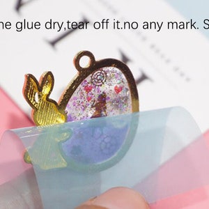 May include: A clear, round, resin pendant with a gold frame and a glittery, purple and pink interior. A gold bunny charm is attached to the frame. A clear, blue protective sheet is being peeled off the pendant.
