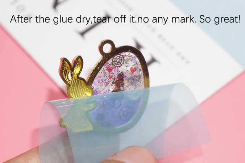 May include: A clear, round, resin pendant with a gold frame and a glittery, purple and pink interior. A gold bunny charm is attached to the frame. A clear, blue protective sheet is being peeled off the pendant.