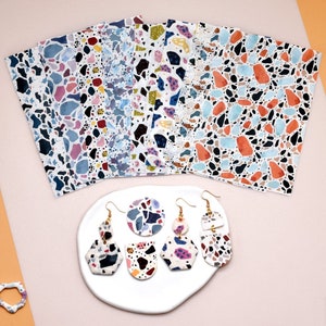 May include: A set of 10 colourful patterned sheets of paper with a terrazzo design. The sheets are arranged in a fan shape. There are four pairs of earrings with a terrazzo design on a white plate.