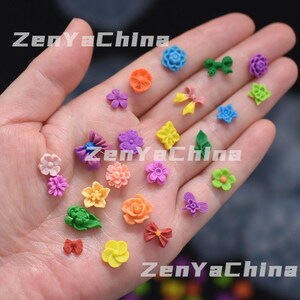 May include: A collection of small, colourful floral and bow-shaped decorations in shades of pink, orange, yellow, green, blue, and purple. The decorations are held in a hand, with the text "ZenYaChina" visible.
