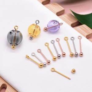 May include: A selection of jewellery-making components, featuring clear, yellow, and purple glass beads with metal detailing. Several metal eye pins in gold, silver, and rose gold are shown, alongside a single gold bead.