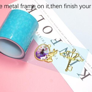 May include: A roll of turquoise blue epoxy resin tape, a gold heart-shaped pendant with a rabbit and a gold key with a crown on top. The text "Put the metal frame on it, then finish your design." is visible in the image.