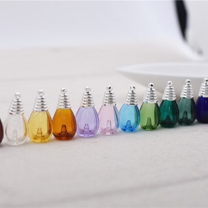 May include: A row of small, colorful glass bottles with silver-colored bulb-shaped tops. The bottles are filled with various shades of liquid, including red, blue, green, and yellow. The bottles are arranged in a line, with a white background.