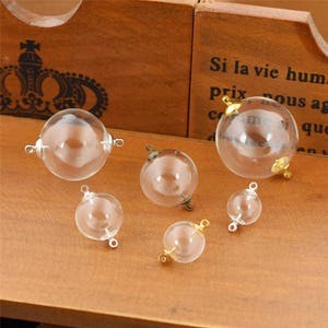 May include: Six clear glass spheres with silver and gold metal loops. The spheres are of varying sizes and are arranged on a wooden surface.