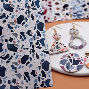 May include: A pair of colourful terrazzo earrings with a triangle and a circle design. The earrings are made of polymer clay and have a white background with blue, pink, and grey speckles.