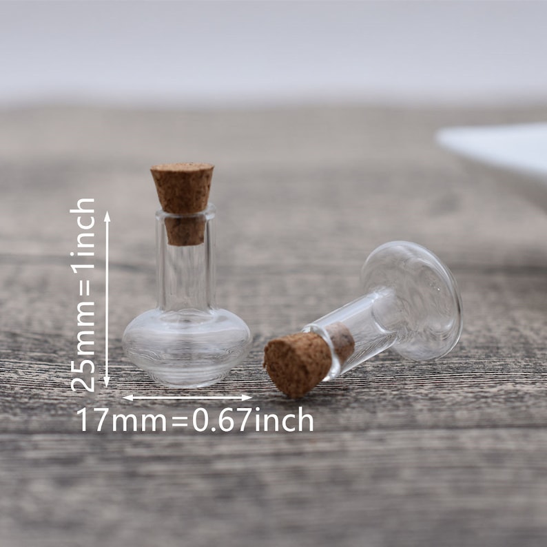May include: Two small clear glass bottles with cork stoppers. The bottles are about 1.7 inches tall and 0.67 inches wide. The bottles are on a wooden surface.