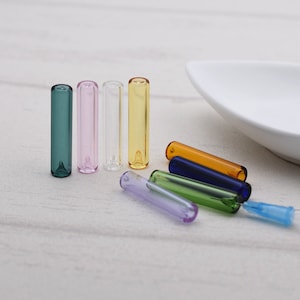 May include: A set of seven glass tubes in various colors, including clear, green, pink, yellow, purple, blue, and orange. The tubes are all approximately the same size and shape, and they are arranged in a semi-circle on a white surface.