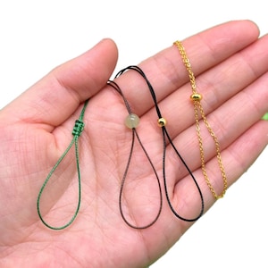 May include: Three different types of phone charms. One is a green cord with a knot at the top and a small bead. Another is a brown cord with a small bead and a knot at the top. The last is a gold chain with two small gold beads.
