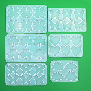 May include: Six silicone molds for making resin jewelry. The molds are arranged in two rows of three. The top row features a clover leaf design, the middle row features a heart design, and the bottom row features a butterfly design. The molds are all clear and have a smooth surface.