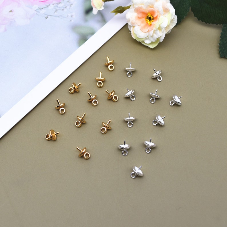 May include: A collection of gold and silver bead caps with a loop on top. The bead caps are arranged in a pattern on a brown surface.