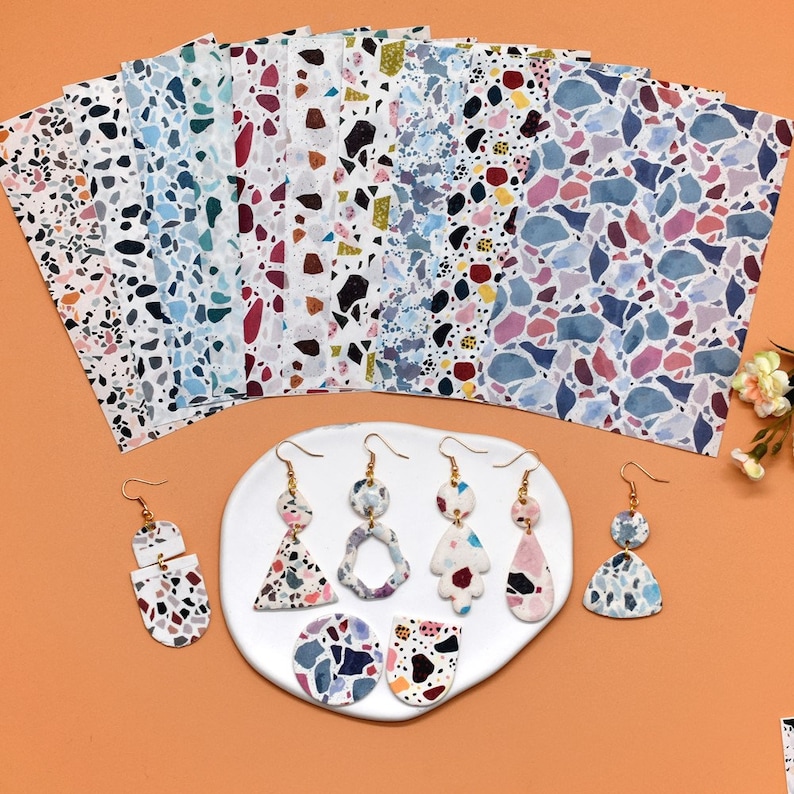 May include: A set of colourful terrazzo patterned paper sheets and a collection of handmade terrazzo earrings with gold accents. The earrings are displayed on a white plate.