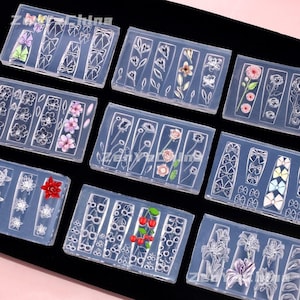 Op de afbeelding: Een collectie rechthoekige siliconen mallen voor nail art, met diverse reliëfmotieven zoals strikjes, bloemen en kersen. Sommige mallen bevatten gekleurde harsaccenten, terwijl andere transparant zijn en ingewikkelde patronen voor nageldecoratie tonen.