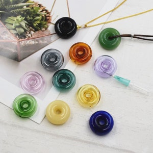 May include: Assortment of 10 colorful glass donut beads, including black, green, purple, amber, gray, light green, light pink, light yellow, and blue. The beads are arranged on a white surface.