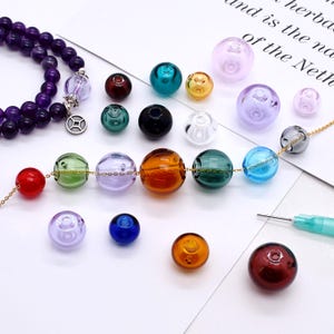 Peut inclure: Un assortiment de perles de verre colorées de différentes tailles et teintes, dont rouge, vert, bleu et orange. Certaines perles sont enfilées sur des chaînes dorées, tandis que d'autres sont en vrac. Un bracelet de perles violet est également visible.